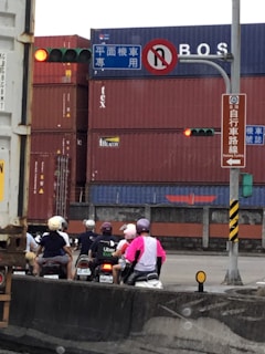 A group of people on scooters waiting at a traffic light near a container yard. Several shipping containers are stacked in the background, and traffic signs are visible overhead. Some riders are wearing helmets with vibrant colors, including pink and purple.