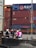 A group of people on scooters waiting at a traffic light near a container yard. Several shipping containers are stacked in the background, and traffic signs are visible overhead. Some riders are wearing helmets with vibrant colors, including pink and purple.