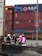 A group of people on scooters waiting at a traffic light near a container yard. Several shipping containers are stacked in the background, and traffic signs are visible overhead. Some riders are wearing helmets with vibrant colors, including pink and purple.
