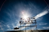 A large metal sign with the words 'Public Market' is against the backdrop of a clear blue sky. The sun is shining brightly behind the sign, creating a lens flare effect. Some clouds are visible on the horizon, and a fish-shaped weather vane is attached to the structure.