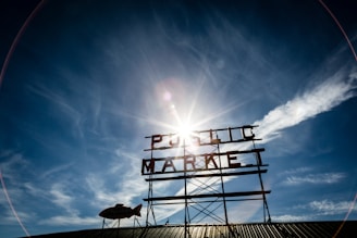 A large metal sign with the words 'Public Market' is against the backdrop of a clear blue sky. The sun is shining brightly behind the sign, creating a lens flare effect. Some clouds are visible on the horizon, and a fish-shaped weather vane is attached to the structure.