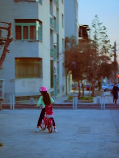 A child wearing a pink helmet rides a pink bicycle along a paved path in an urban setting. Apartment buildings with balconies and windows serve as the backdrop. Trees and parked cars are visible in the background.