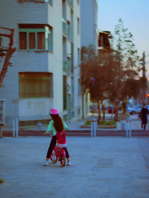 A child wearing a pink helmet rides a pink bicycle along a paved path in an urban setting. Apartment buildings with balconies and windows serve as the backdrop. Trees and parked cars are visible in the background.