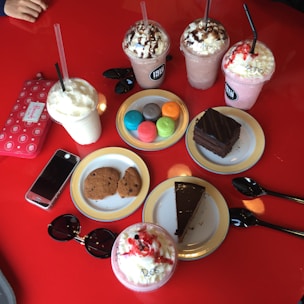 An assortment of colorful desserts and drinks on a bright red table, including various milkshakes topped with whipped cream and chocolate syrup, colorful macarons, cookies, slices of chocolate cake, a smartphone, sunglasses, and a wallet.