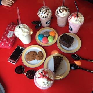 An assortment of colorful desserts and drinks on a bright red table, including various milkshakes topped with whipped cream and chocolate syrup, colorful macarons, cookies, slices of chocolate cake, a smartphone, sunglasses, and a wallet.
