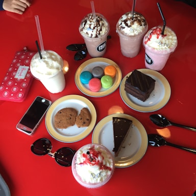 An assortment of colorful desserts and drinks on a bright red table, including various milkshakes topped with whipped cream and chocolate syrup, colorful macarons, cookies, slices of chocolate cake, a smartphone, sunglasses, and a wallet.