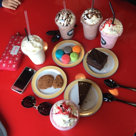 An assortment of colorful desserts and drinks on a bright red table, including various milkshakes topped with whipped cream and chocolate syrup, colorful macarons, cookies, slices of chocolate cake, a smartphone, sunglasses, and a wallet.