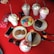 An assortment of colorful desserts and drinks on a bright red table, including various milkshakes topped with whipped cream and chocolate syrup, colorful macarons, cookies, slices of chocolate cake, a smartphone, sunglasses, and a wallet.