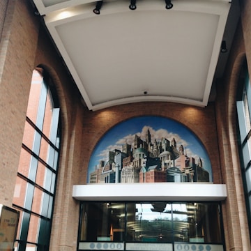 A large, arched mural depicting a cityscape adorns the interior of a building. The mural features a blend of historical and modern architecture with diverse building styles against a partly cloudy sky. The surrounding interior space is characterized by high, brick walls and large, vertical windows that allow natural light to flood the area.