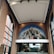A large, arched mural depicting a cityscape adorns the interior of a building. The mural features a blend of historical and modern architecture with diverse building styles against a partly cloudy sky. The surrounding interior space is characterized by high, brick walls and large, vertical windows that allow natural light to flood the area.