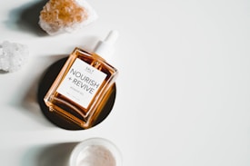 A bottle of rosehip oil labeled 'Nourish + Revive' is placed on a dark circular surface. Surrounding the bottle are two pieces of raw crystals and a small open container with a similar color to the oil.