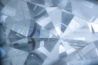 A close-up view of a cut and polished gemstone with a complex array of reflective facets that refract light, creating a sparkling effect. The facets are sharply defined, intersecting at various angles, and the overall texture appears smooth and glass-like.