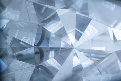 A close-up view of a cut and polished gemstone with a complex array of reflective facets that refract light, creating a sparkling effect. The facets are sharply defined, intersecting at various angles, and the overall texture appears smooth and glass-like.