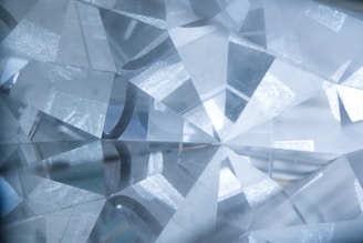 A close-up view of a cut and polished gemstone with a complex array of reflective facets that refract light, creating a sparkling effect. The facets are sharply defined, intersecting at various angles, and the overall texture appears smooth and glass-like.