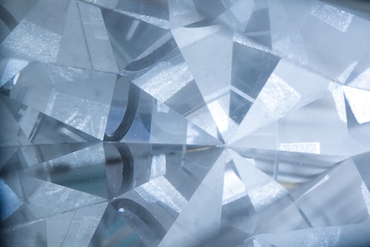 A close-up view of a cut and polished gemstone with a complex array of reflective facets that refract light, creating a sparkling effect. The facets are sharply defined, intersecting at various angles, and the overall texture appears smooth and glass-like.