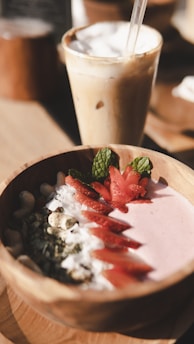 A wooden bowl filled with a pink smoothie, garnished with fresh sliced strawberries, mint leaves, seeds, and nuts, sits in the foreground. Behind it, a glass of iced coffee with a straw and a layer of frothy milk on top adds to the inviting breakfast or snack scene.