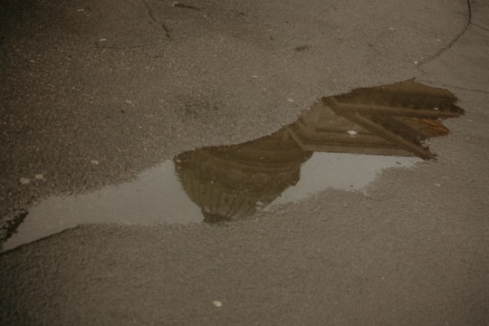 A puddle on an asphalt surface reflects part of a building, possibly a domed structure, in murky water. The pavement appears worn with visible cracks.