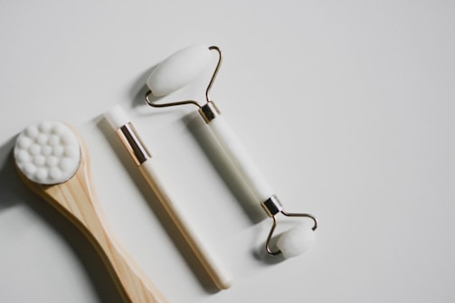 Three beauty tools are arranged on a white surface. A wooden-handled brush with white bristles sits on the left, a white-handled brush with a small, rounded brush head is in the middle, and a white jade roller with metal accents is on the right. The tools appear to be clean and unused.