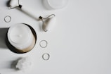 A white candle in a jar, a facial roller, several golden rings, and a cluster of white crystals are arranged on a white background.