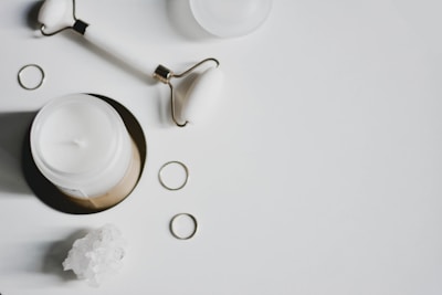A white candle in a jar, a facial roller, several golden rings, and a cluster of white crystals are arranged on a white background.
