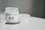 A white candle jar labeled 'THE RELATABLE CANDLE CO. No.6 WE BE CHILLIN' is placed on a white surface. The candle is lime, basil, and orange scented. There is a small cluster of white crystals on the left side.