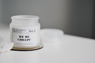 A white candle jar labeled 'THE RELATABLE CANDLE CO. No.6 WE BE CHILLIN' is placed on a white surface. The candle is lime, basil, and orange scented. There is a small cluster of white crystals on the left side.