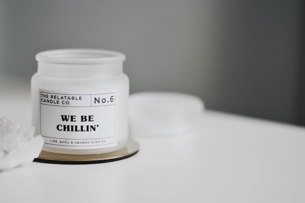 A white candle jar labeled 'THE RELATABLE CANDLE CO. No.6 WE BE CHILLIN' is placed on a white surface. The candle is lime, basil, and orange scented. There is a small cluster of white crystals on the left side.