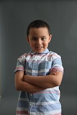 A young child stands confidently with arms crossed, wearing a striped shirt with pastel shades against a plain, dark background. The child has short dark hair and is smiling gently.
