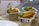 Three foil containers are filled with what appears to be a savory dish topped with crispy fried sticks, garnished with fresh green herbs and small red tomato pieces. The containers are placed on a wooden board, emphasizing a rustic presentation.