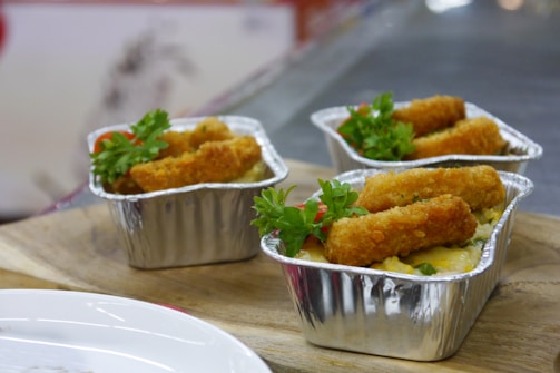 Three foil containers are filled with what appears to be a savory dish topped with crispy fried sticks, garnished with fresh green herbs and small red tomato pieces. The containers are placed on a wooden board, emphasizing a rustic presentation.