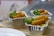 Three foil containers are filled with what appears to be a savory dish topped with crispy fried sticks, garnished with fresh green herbs and small red tomato pieces. The containers are placed on a wooden board, emphasizing a rustic presentation.