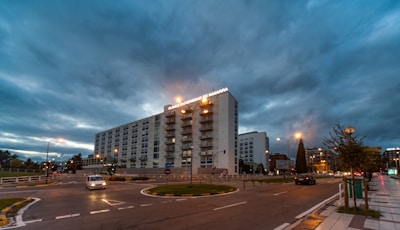 A large multi-story building dominates the scene, with signage indicating it is a university clinic. The sky is overcast with dramatic clouds, giving the atmosphere a moody feel. Several cars are navigating the roundabout in front of the building, and streetlights are illuminated, suggesting a time around dusk or dawn. Trees and urban infrastructure such as roads and sidewalks are visible.