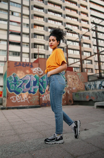 Urban street scene with a young adult in a muted gold nisqala tee, walking confidently past graffiti