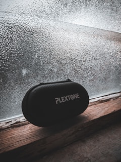 A black, textured carrying case with the brand name 'PlexTone' in white text sits on a wooden windowsill. The background features a frosted glass window with a textured pattern, through which diffuse light filters, creating a subdued, muted atmosphere.