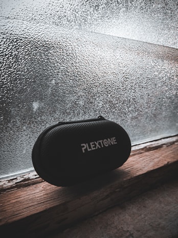 A black, textured carrying case with the brand name 'PlexTone' in white text sits on a wooden windowsill. The background features a frosted glass window with a textured pattern, through which diffuse light filters, creating a subdued, muted atmosphere.