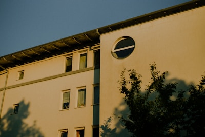 A residential building with a modern architectural design featuring rectangular windows and a distinctive circular window on the upper floor. The facade is illuminated by warm sunlight, casting shadows of nearby trees across the wall.