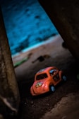 A small orange toy car with floral decorations is placed on a sandy surface between two large structures. The background features a blurred mix of earthy and blue tones, suggesting an outdoor or creative setting.