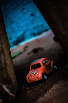A small orange toy car with floral decorations is placed on a sandy surface between two large structures. The background features a blurred mix of earthy and blue tones, suggesting an outdoor or creative setting.