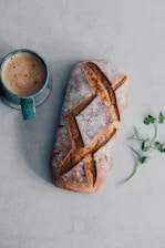 A loaf of rustic bread with a golden crust is placed beside a green ceramic mug filled with coffee. A small sprig of green leaves lies nearby on a light gray surface.