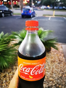 A hand holds a Coca-Cola bottle with an orange vanilla flavor label prominently displayed. The background features a parking lot with several cars and blurred greenery.