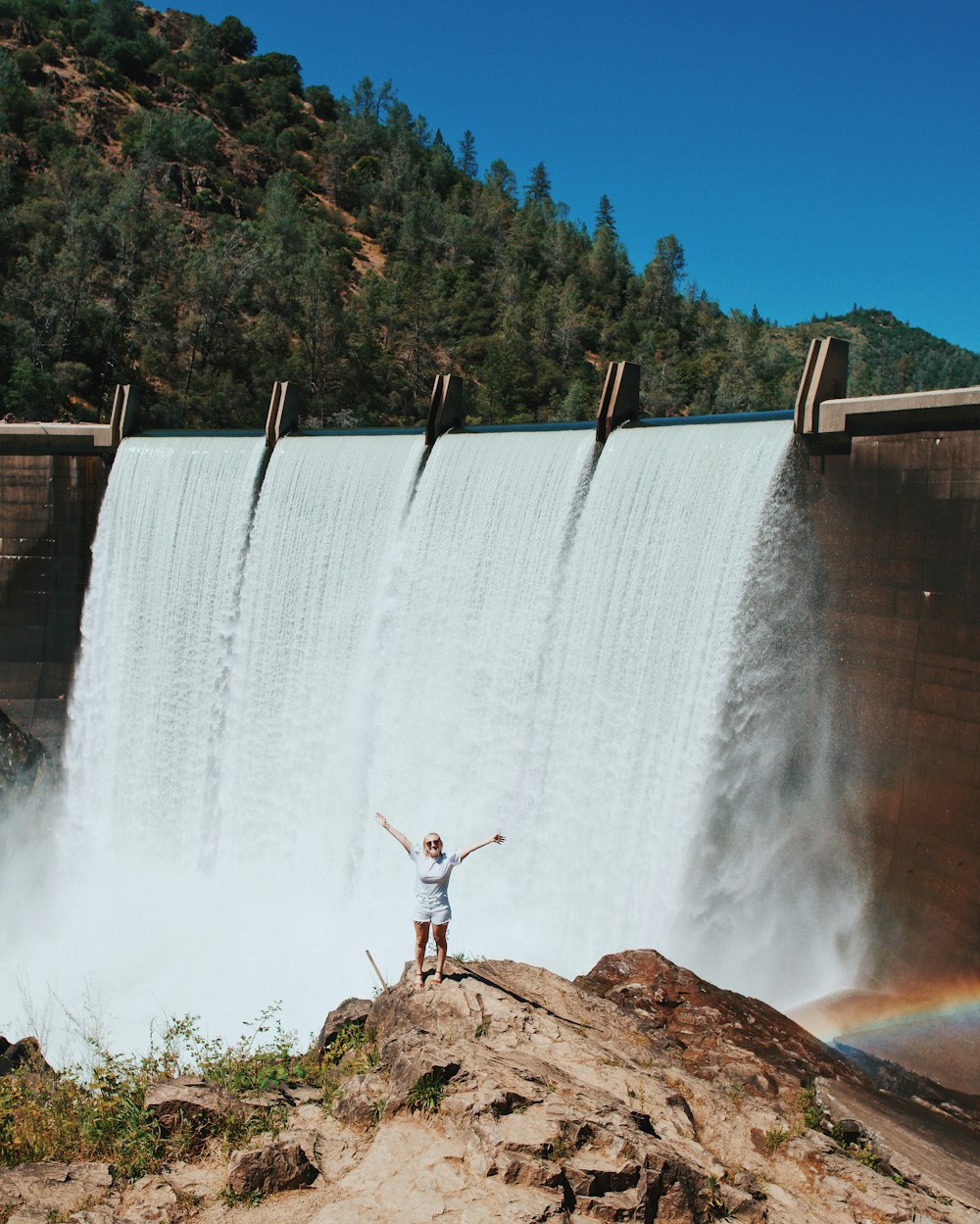 man in red shirt standing on brown rock near waterfalls during daytime