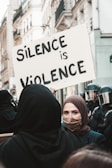 A protest scene in an urban area. A woman wearing a headscarf and face mask holds a large sign that reads 'Silence is Violence.' In the background, there are police officers wearing helmets and face shields. The photo is taken on a street lined with tall buildings.