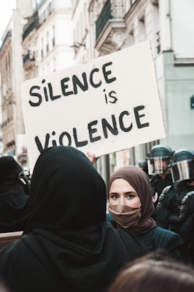 A protest scene in an urban area. A woman wearing a headscarf and face mask holds a large sign that reads 'Silence is Violence.' In the background, there are police officers wearing helmets and face shields. The photo is taken on a street lined with tall buildings.