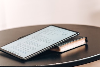 A tablet displaying a document is placed on top of a closed book. Both items rest on a round black table. The document on the tablet screen appears to be text-based, possibly an article or notes.
