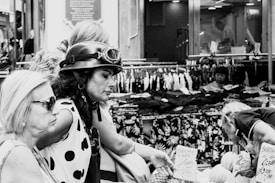 A black and white street market scene with several people engaged in shopping activities. One person is wearing a helmet with goggles while others appear to be examining goods. Various clothing items are displayed in the background.