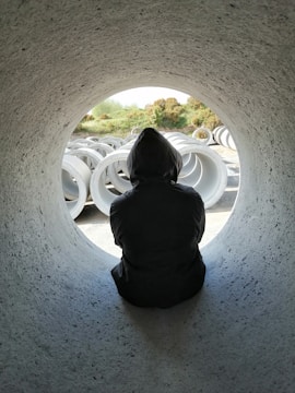 A person wearing a hoodie sits inside a large concrete pipe, looking outward. Outside, there is a collection of similar pipes stacked in an open area with greenery and bushes visible in the background.