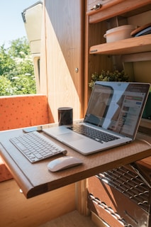 A compact workspace within a small wooden room, possibly a van or camper, featuring a wooden table with a laptop and a keyboard. The table is foldable and the room also contains some shelves with items like bowls and plants. A mug is placed near the laptop, which displays a website. Natural light enters the space, creating a warm and cozy atmosphere.