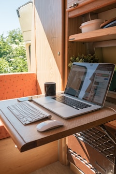 A compact workspace within a small wooden room, possibly a van or camper, featuring a wooden table with a laptop and a keyboard. The table is foldable and the room also contains some shelves with items like bowls and plants. A mug is placed near the laptop, which displays a website. Natural light enters the space, creating a warm and cozy atmosphere.