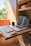 A compact workspace within a small wooden room, possibly a van or camper, featuring a wooden table with a laptop and a keyboard. The table is foldable and the room also contains some shelves with items like bowls and plants. A mug is placed near the laptop, which displays a website. Natural light enters the space, creating a warm and cozy atmosphere.