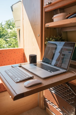A compact workspace within a small wooden room, possibly a van or camper, featuring a wooden table with a laptop and a keyboard. The table is foldable and the room also contains some shelves with items like bowls and plants. A mug is placed near the laptop, which displays a website. Natural light enters the space, creating a warm and cozy atmosphere.